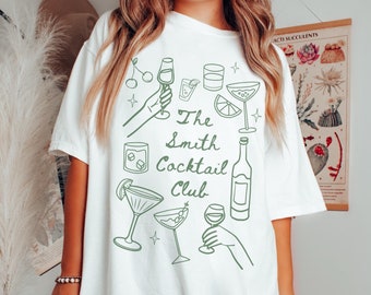 Op de afbeelding: Een wit t-shirt met een groene lijntekening van diverse cocktailglazen, een fles en handen die drankjes vasthouden. De tekst "The Smith Cocktail Club" staat in het midden gedrukt in een groen scriptlettertype, omringd door kleine sterren.