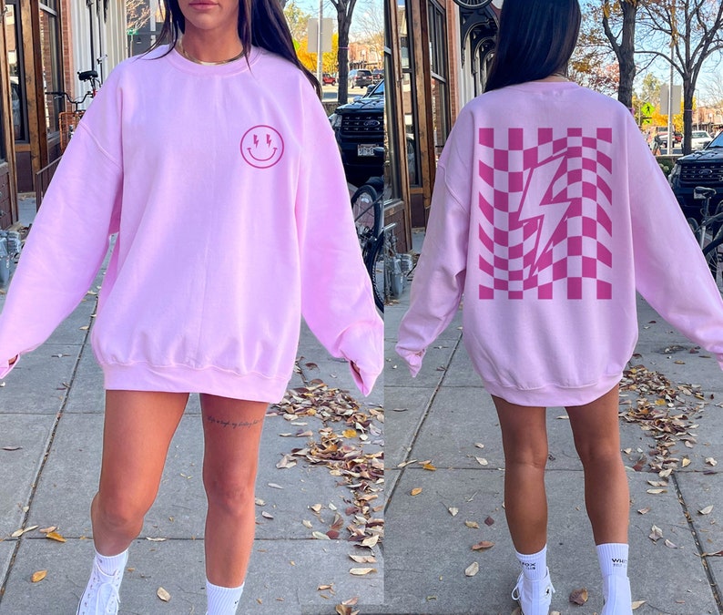 May include: A light pink crewneck sweatshirt featuring a small dark pink smiley face with lightning bolt eyes on the front left chest and a large dark pink wavy checkerboard pattern with a central lightning bolt graphic on the back.