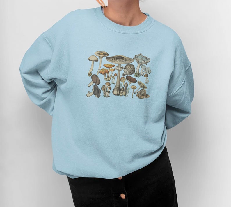 May include: A light blue crewneck sweatshirt featuring a central graphic print of various mushrooms in shades of brown, tan, and muted orange. The long-sleeved pullover has a ribbed collar, cuffs, and hem, providing a standard fit and finish.