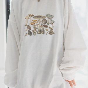 May include: A white long-sleeve crewneck sweatshirt featuring a detailed, multi-colored graphic print of various mushroom species in earthy tones of brown, tan, and muted blue, arranged in a cluster across the center of the chest.