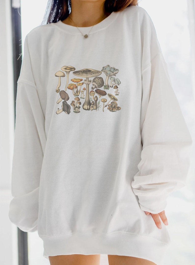 May include: A white long-sleeve crewneck sweatshirt featuring a detailed, multi-colored graphic print of various mushroom species in earthy tones of brown, tan, and muted blue, arranged in a cluster across the center of the chest.