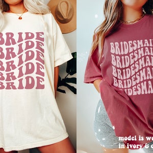 May include: An ivory t-shirt with the word BRIDE printed in a repeating wavy dark pink font and a crimson t-shirt with the word BRIDESMAID printed in a repeating wavy light pink font. The text on the crimson shirt states model is wearing size XL.