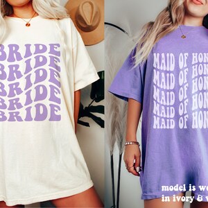 May include: Two short-sleeve t-shirts. The left shirt is ivory with the word BRIDE repeated in a wavy purple font. The right shirt is violet with the text MAID OF HONOR repeated in a wavy white font. The text states the model is wearing size XL.