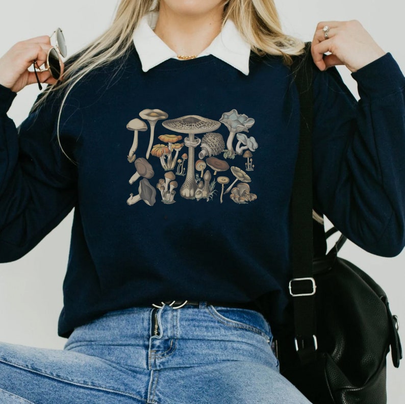 May include: A navy blue long-sleeve sweatshirt featuring a detailed graphic print of various mushrooms in earthy tones of brown, tan, and grey. The sweatshirt has a white collared neckline and is worn with blue denim jeans and a black shoulder bag.