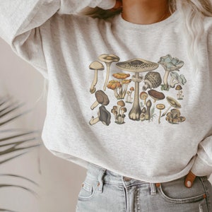 May include: A light gray heathered crewneck sweatshirt featuring a detailed graphic print of various mushrooms in earthy tones including brown, tan, orange, and muted green. The illustration displays a collection of fungi with diverse cap shapes.