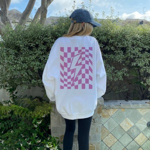 May include: A white long-sleeve sweatshirt featuring a large, centered graphic on the back. The graphic consists of a pink and white distorted, wavy checkerboard pattern with a solid pink lightning bolt shape overlaid in the center.