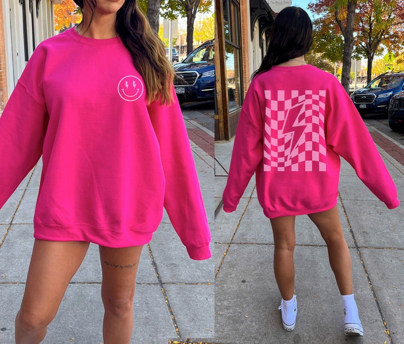 May include: A bright hot pink crewneck sweatshirt featuring a small white smiley face with lightning bolt eyes on the left chest and a large, distorted checkerboard pattern with a central white lightning bolt graphic on the back.