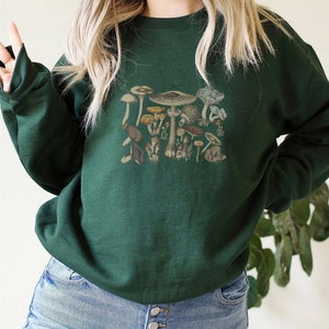 May include: A dark forest green crewneck sweatshirt featuring a central graphic print of various mushrooms in earthy tones of brown, tan, and muted orange. The long-sleeved pullover has a ribbed neckline, cuffs, and hem, providing a classic fit.