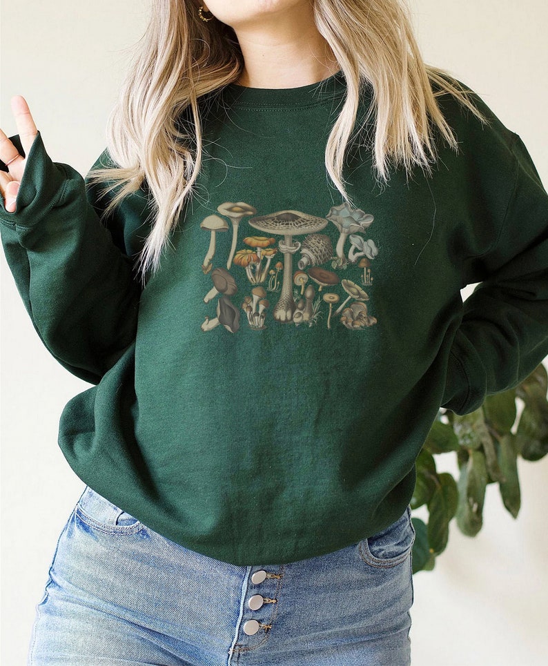 May include: A dark forest green crewneck sweatshirt featuring a central graphic print of various mushrooms in earthy tones of brown, tan, and muted orange. The long-sleeved pullover has a ribbed neckline, cuffs, and hem, providing a classic fit.