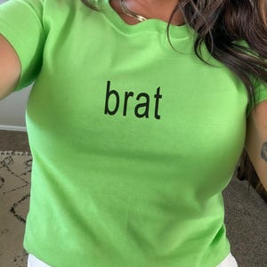 May include: A bright lime green short-sleeved t-shirt featuring the word brat printed in a simple black sans-serif font across the center of the chest. The garment has a classic crew neckline and a fitted silhouette made of a solid-colored fabric.