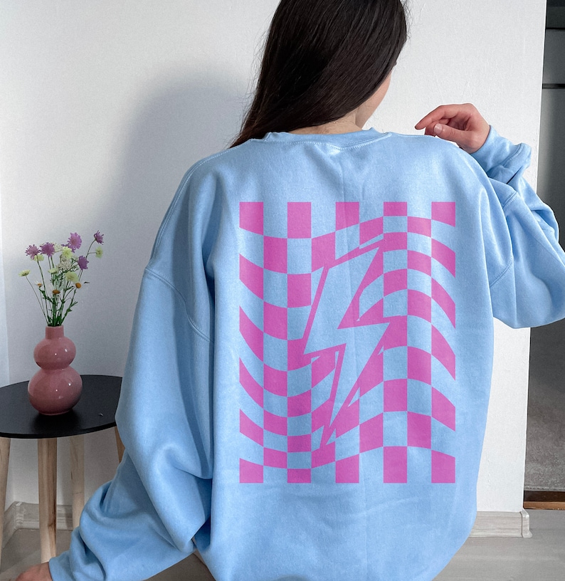 May include: A light blue crewneck sweatshirt featuring a large, centered graphic on the back. The design consists of a pink and light blue wavy checkerboard pattern with a prominent white lightning bolt shape overlaid in the center of the grid.