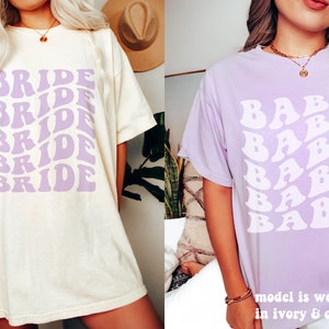 May include: An ivory t-shirt with the word BRIDE printed in a repeating, wavy purple font, and a light purple t-shirt with the word BABE printed in a repeating, wavy white font. The text on the image states model is wearing size XL in ivory & orchid.