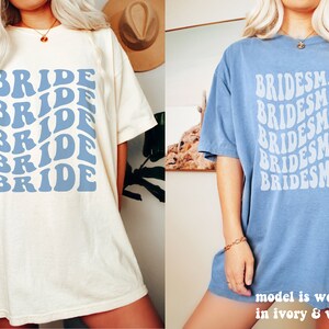 May include: Two short-sleeve crew neck t-shirts. The left shirt is ivory with blue text reading BRIDE in a repeating wavy pattern. The right shirt is washed denim blue with white text reading BRIDESMAID in a repeating wavy pattern.