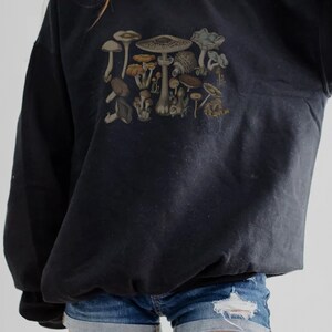 May include: A black long-sleeve sweatshirt featuring a detailed graphic print of various mushrooms in earthy tones of brown, tan, and muted green. The sweatshirt is paired with blue denim shorts that have a distressed, frayed hem detail.