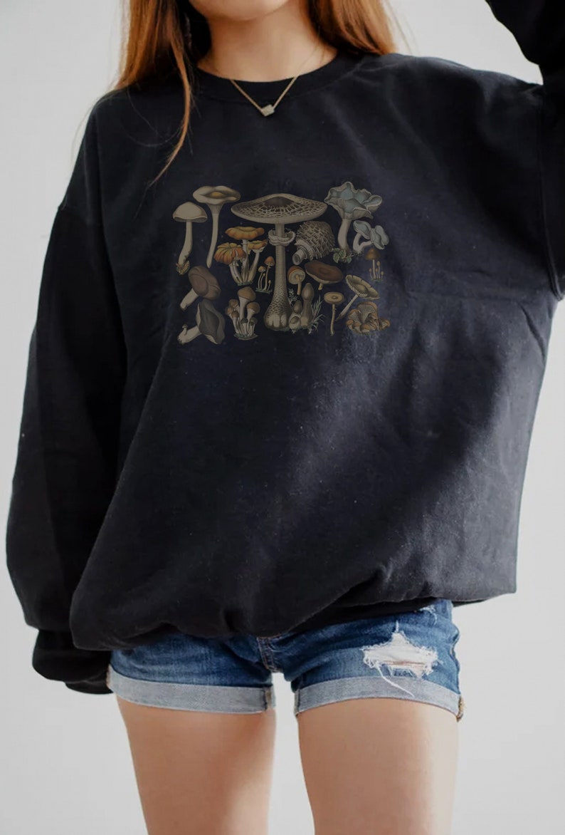 May include: A black long-sleeve sweatshirt featuring a detailed graphic print of various mushrooms in earthy tones of brown, tan, and muted green. The sweatshirt is paired with blue denim shorts that have a distressed, frayed hem detail.