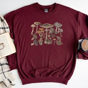 May include: A maroon crewneck sweatshirt featuring a central graphic print of various mushrooms in shades of brown, tan, and cream. The long-sleeved garment has a ribbed collar, cuffs, and hem, showcasing a detailed botanical illustration of fungi.