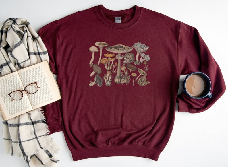 May include: A maroon crewneck sweatshirt featuring a central graphic print of various mushrooms in shades of brown, tan, and cream. The long-sleeved garment has a ribbed collar, cuffs, and hem, showcasing a detailed botanical illustration of fungi.