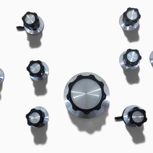 May include: A collection of ten circular control knobs featuring a brushed silver metal center and a black fluted outer rim. The set includes one large knob and nine smaller knobs, all designed with a consistent industrial aesthetic and finish.