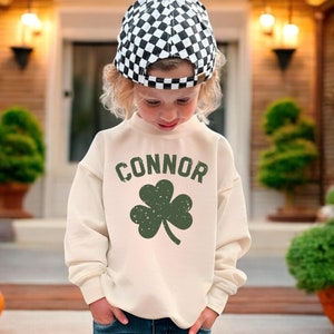 May include: A child wearing a white sweatshirt with the name "CONNOR" and a green shamrock printed on it. The child is also wearing a black and white checkered hat and blue jeans.