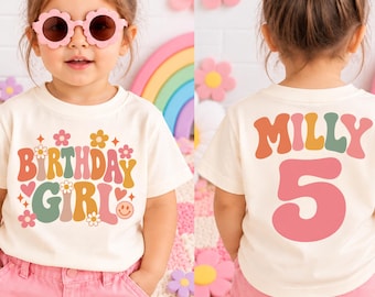 May include: A cream-coloured short-sleeve t-shirt featuring the text "BIRTHDAY GIRL" with colourful flowers, hearts, and a smiley face on the front, and the name "MILLY" above the number "5" in large, bold, multi-coloured lettering on the back.