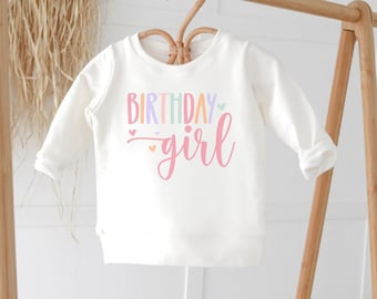 May include: White long-sleeved shirt with the text "birthday girl" in rainbow colors with pink hearts.