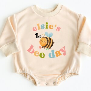 May include: A cream-colored long-sleeved onesie with a bee and flower design. The text on the onesie reads "elsie's 1st bee day" in colorful letters.