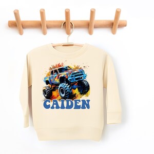 May include: A cream colored sweatshirt with a colorful monster truck graphic and the name "CAIDEN" in blue letters.