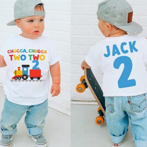 May include: A white toddler t-shirt with a colorful train graphic and the text "CHUGGA, CHUGGA TWO TWO." The back of the shirt has the name "JACK" and the number "2" in blue. The child is wearing a gray cap and blue jeans.