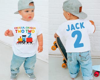 May include: A white toddler t-shirt with a colorful train graphic and the text "CHUGGA, CHUGGA TWO TWO." The back of the shirt has the name "JACK" and the number "2" in blue. The child is wearing a gray cap and blue jeans.