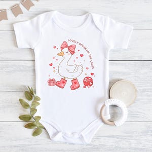 May include: White baby onesie with a cartoon goose wearing a pink bow and boots. The goose is surrounded by hearts and the text "LOVELY GOOSE ON THE LOOSE". A gift box and a heart-shaped container are also featured.