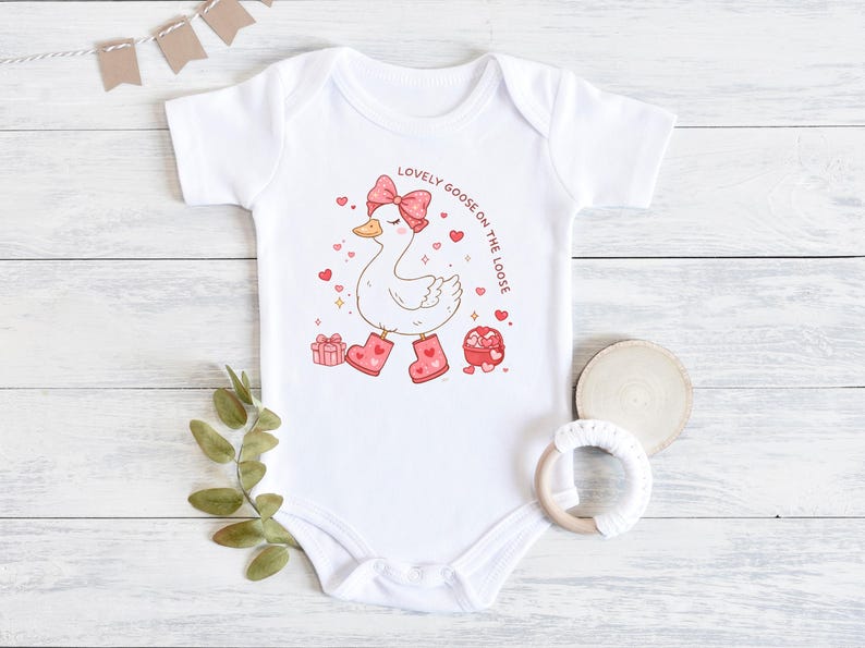 May include: White baby onesie with a cartoon goose wearing a pink bow and boots. The goose is surrounded by hearts and the text "LOVELY GOOSE ON THE LOOSE". A gift box and a heart-shaped container are also featured.