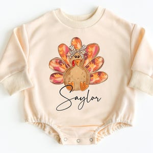 May include: A cream-colored long-sleeved bodysuit with a graphic of a turkey wearing a bow. The turkey has orange, red, and brown feathers. The text "Saylor" is printed below the turkey.