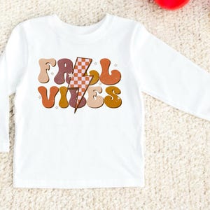 May include: White long-sleeve shirt with a retro-style graphic that says "Fall Vibes" in orange, brown, and yellow letters with a checkered lightning bolt in the middle.