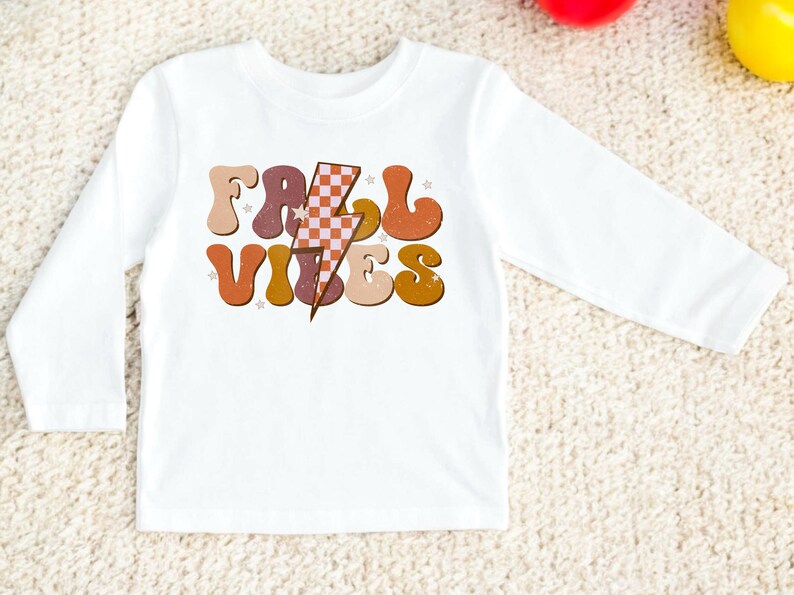 May include: White long-sleeve shirt with a retro-style graphic that says "Fall Vibes" in orange, brown, and yellow letters with a checkered lightning bolt in the middle.
