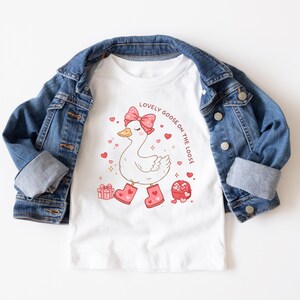 May include: White t-shirt with a cartoon goose wearing a pink bow and boots, surrounded by hearts and gifts. The text "LOVELY GOOSE ON THE LOOSE" is printed above the goose. The shirt is paired with a blue denim jacket.