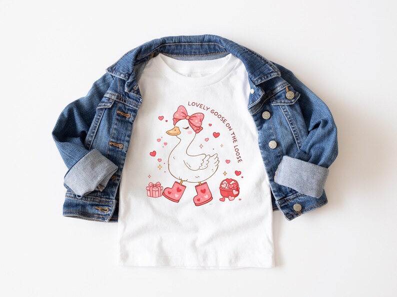 May include: White t-shirt with a cartoon goose wearing a pink bow and boots, surrounded by hearts and gifts. The text "LOVELY GOOSE ON THE LOOSE" is printed above the goose. The shirt is paired with a blue denim jacket.