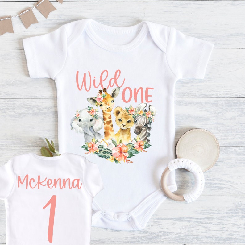 May include: White onesie with a pink 'Wild One' graphic featuring a giraffe, lion, zebra, and elephant with floral accents. The back of the onesie has the name 'Mckenna' and the number '1' in pink.