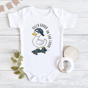 May include: White baby bodysuit featuring a cartoon duck in a checkered cap and shoes, skateboarding. The text "Silly Goose on the Loose" is printed around the duck. A playful design for babies.