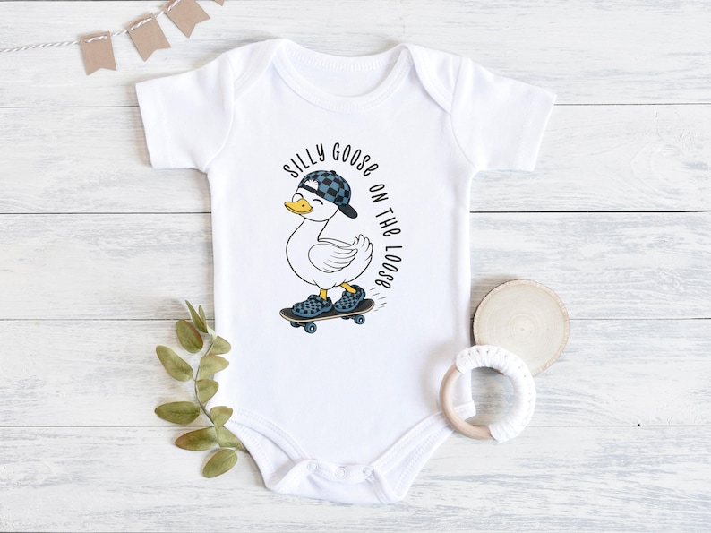 May include: White baby bodysuit featuring a cartoon duck in a checkered cap and shoes, skateboarding. The text "Silly Goose on the Loose" is printed around the duck. A playful design for babies.