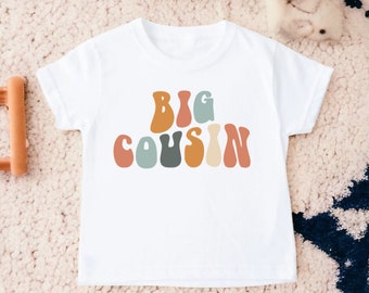 May include: White t-shirt with the words "BIG COUSIN" in colorful, retro-style lettering. The text is in shades of orange, blue, green, and beige. The shirt is likely for a child.