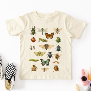 May include: Cream-colored short-sleeved t-shirt featuring a colorful insect illustration. The design includes various bugs like butterflies, bees, and beetles. A pair of black and white checkered slip-on shoes are on the left.