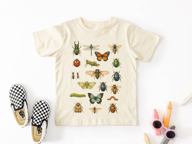 May include: Cream-colored short-sleeved t-shirt featuring a colorful insect illustration. The design includes various bugs like butterflies, bees, and beetles. A pair of black and white checkered slip-on shoes are on the left.