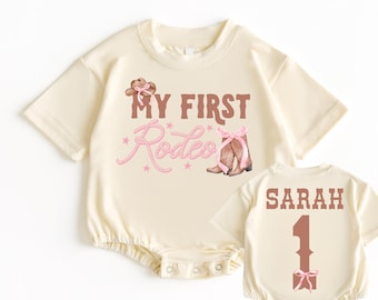 Peut inclure: Un body à manches courtes de couleur crème avec le texte "My First Rodeo" en lettres brunes et des accents roses. Une paire de bottes de cow-boy roses et brunes est représentée sous le texte. Le dos du body porte le nom "SARAH" et le numéro "1" en lettres brunes avec un nœud rose.