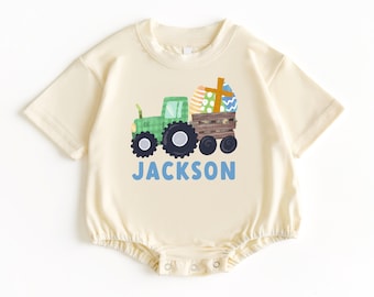 May include: A cream coloured short-sleeved bodysuit with a green tractor carrying Easter eggs and a cross. The text "JACKSON" is printed in blue below the tractor.