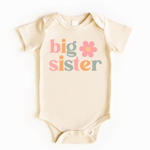 Puede incluir: Body de beb&eacute; color crema con las palabras "big sister" en rosa, naranja y verde, con un gr&aacute;fico de flor rosa. El body est&aacute; hecho de material suave y tiene mangas cortas.