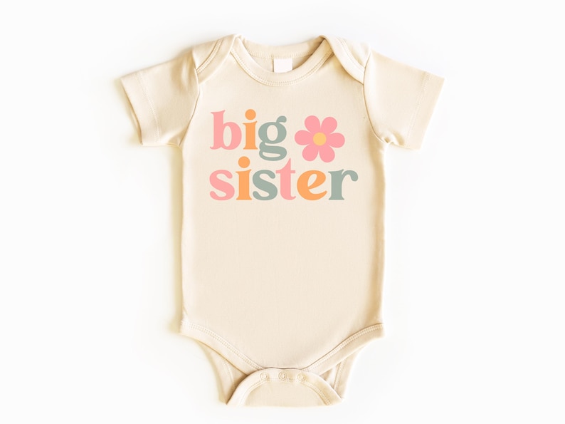 Puede incluir: Body de beb&eacute; color crema con las palabras "big sister" en rosa, naranja y verde, con un gr&aacute;fico de flor rosa. El body est&aacute; hecho de material suave y tiene mangas cortas.