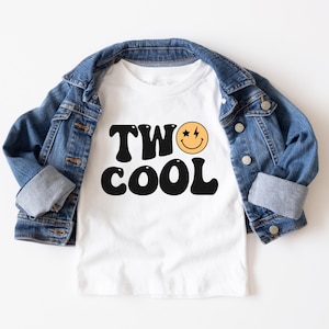 May include: White t-shirt with black text that says "TWO COOL" and a yellow smiley face with stars and a lightning bolt. The shirt is on top of a blue denim jacket.