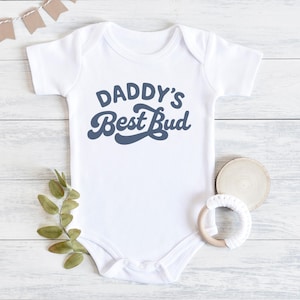 May include: White baby onesie with the text "DADDY'S Best Bud" in a retro blue font. The onesie is laid on a white wooden surface with a sprig of greenery, a wooden disc, and a baby teether.