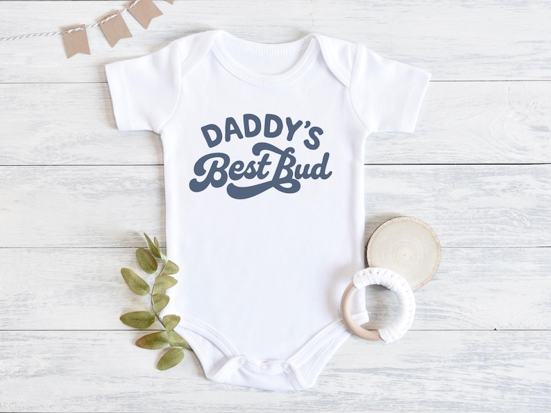 May include: White baby onesie with the text "DADDY'S Best Bud" in a retro blue font. The onesie is laid on a white wooden surface with a sprig of greenery, a wooden disc, and a baby teether.