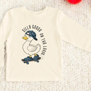 May include: A cream-coloured long-sleeved shirt with a cartoon duck on a skateboard, wearing a chequered cap and shoes. The text "SILLY GOOSE ON THE LOOSE" is printed around the duck. The shirt is made of a soft material.